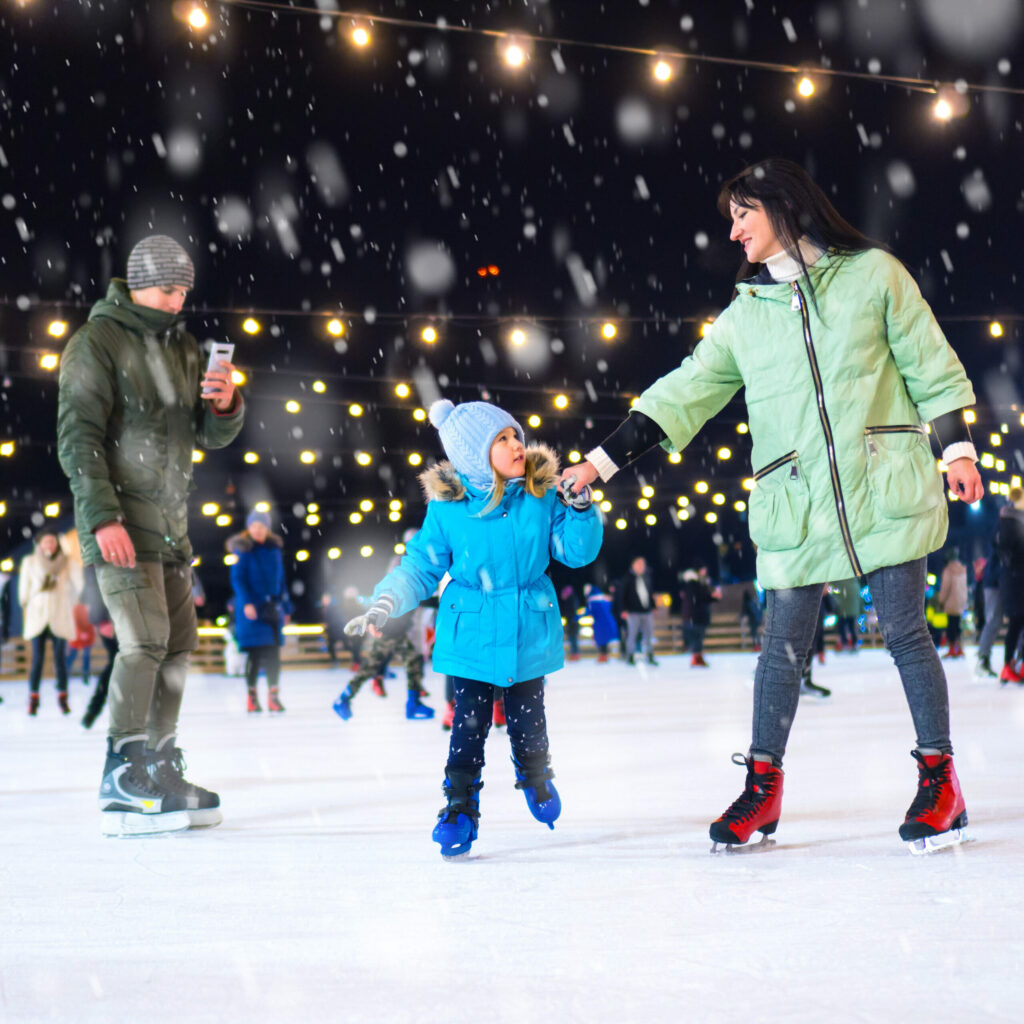 A mom and daughter ice skating while the father records on his phone.