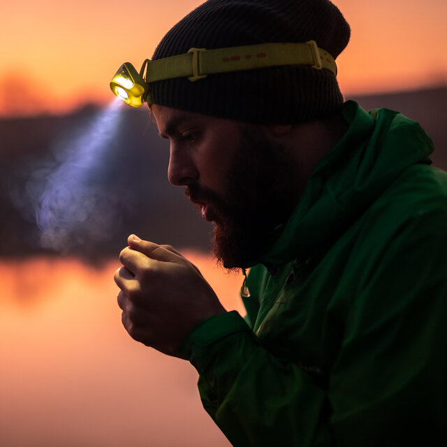 Side view of bearded male traveler in warm clothing and glowing headlamp warming up hands with breath while standing against blurred lake, in nature at sunset time