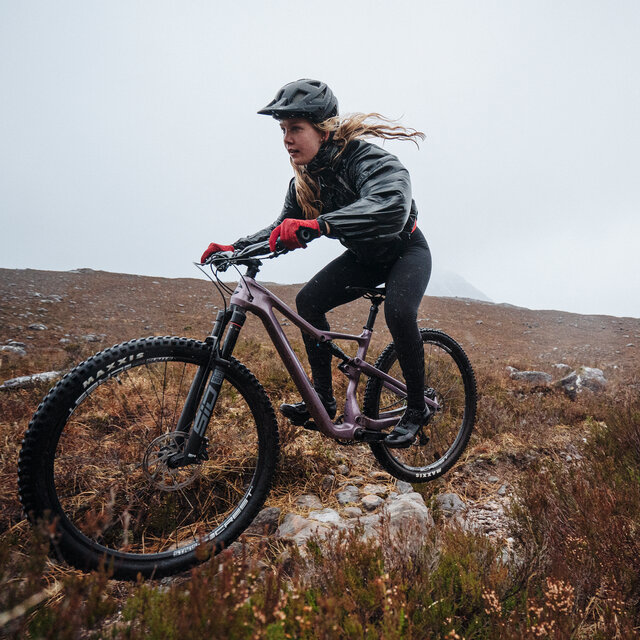 A female rider rides a Cannondale Scalpel Mountain Bike over some rocks.
