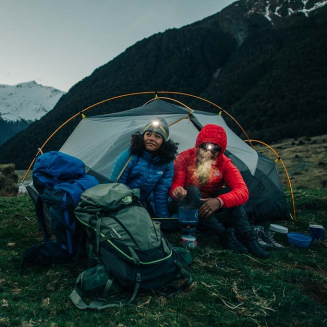 Two backpackers sitting next to a tent and compact stove in a valley.