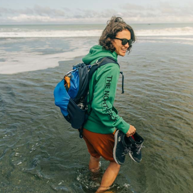 Woman smiling as she holds her shoes and wades through knee-high water.