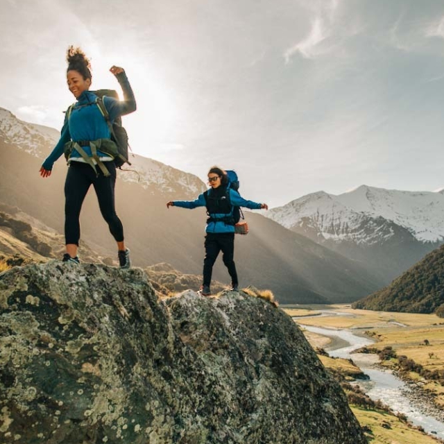 Two women balancing as they backpack over large boulders.