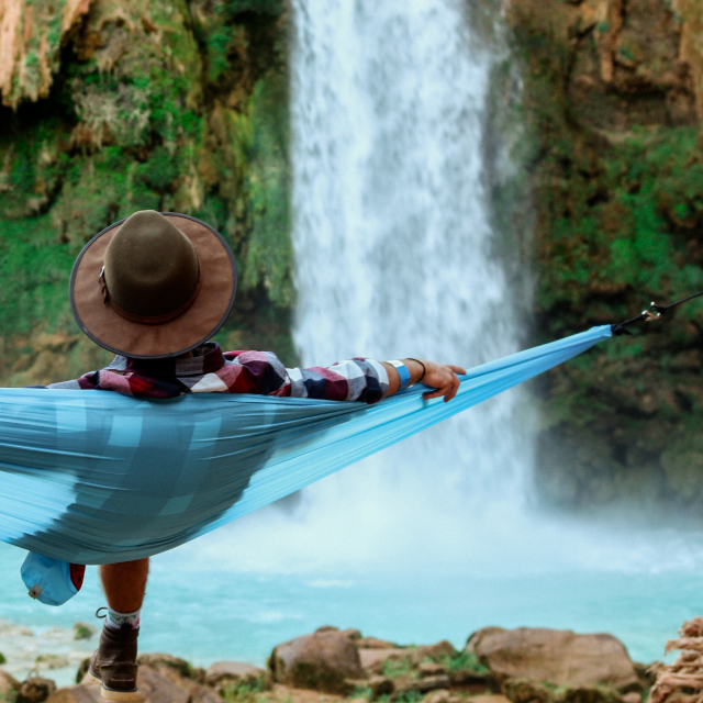 A person relaxing in a thin teal hammock in front of a water fall. The fabric of the hammock body is bunched together where it connects of the hammock straps.