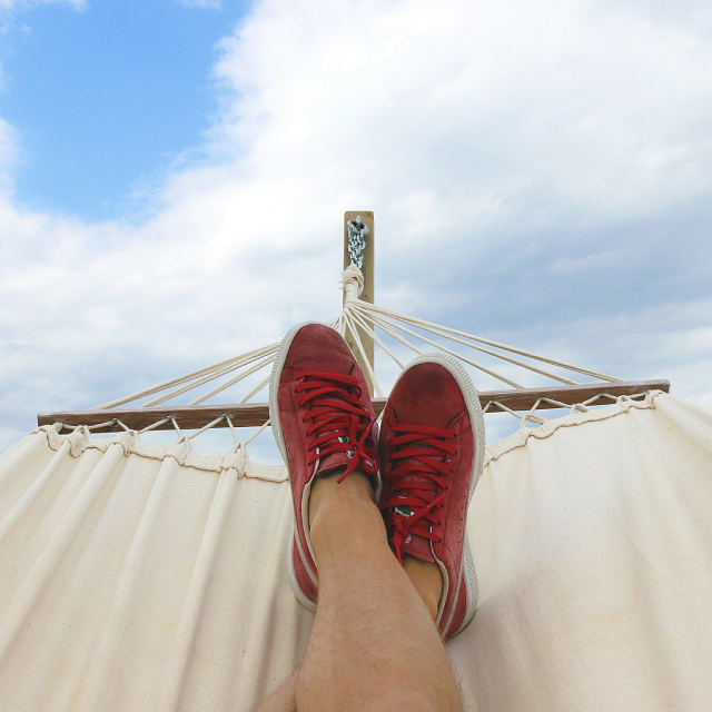 A person wearing red tennis shoes relaxing in a white cotton hammock. The end of the hammock has a wooden bar which prevents the fabric from curling up.