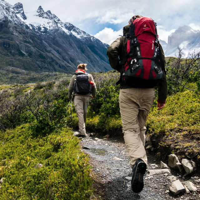 Two backpackers hiking down a scrubland trail towards mountains.