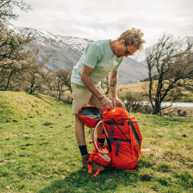 Man adjusting a hiking backpack on a grassy hill.