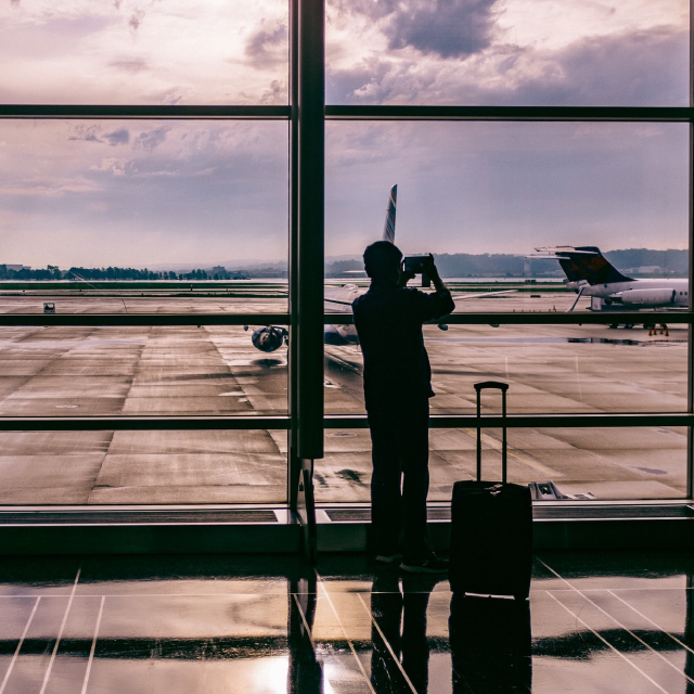 Silhouette of person with carry-on luggage looking out the windows of an airport terminal.
