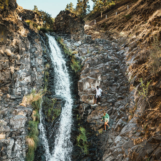 Two people traversing down the rocks beside a sheer waterfall.