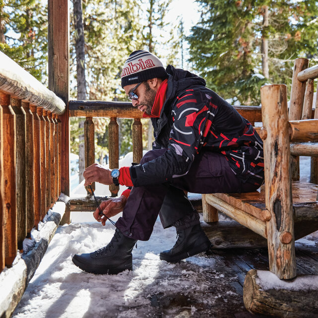A man on a wooden bench, lacing up his boots in the snow.