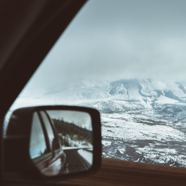 A rearview mirror shows a car driving down an empty road. It is contrasted with a towering, icy hill looming in the distance.