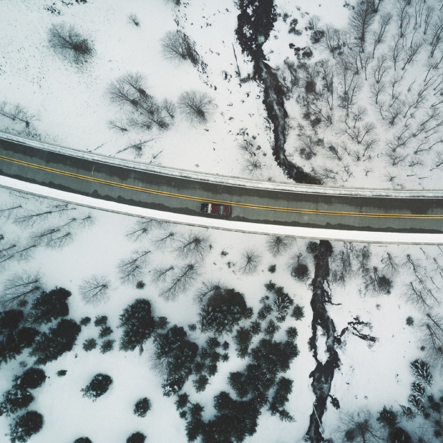 An overhead view of a car as it travels down a two-lane road. The road is surrounded by sparse trees peeking through a blanket of white snow.
