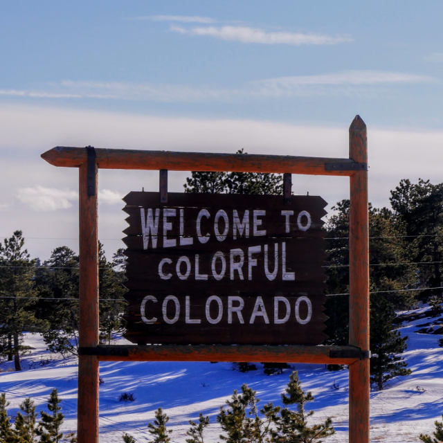 A wooden sign reading “Welcome to colorful Colorado.” Snow blankets the ground and trees in the background.